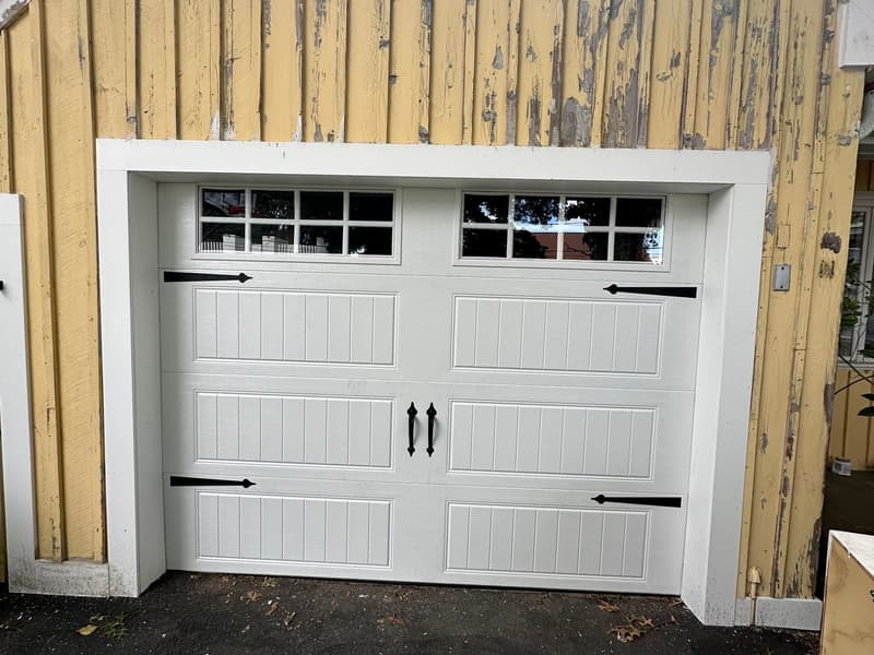 White carriage-style garage door with decorative hardware on older yellow home